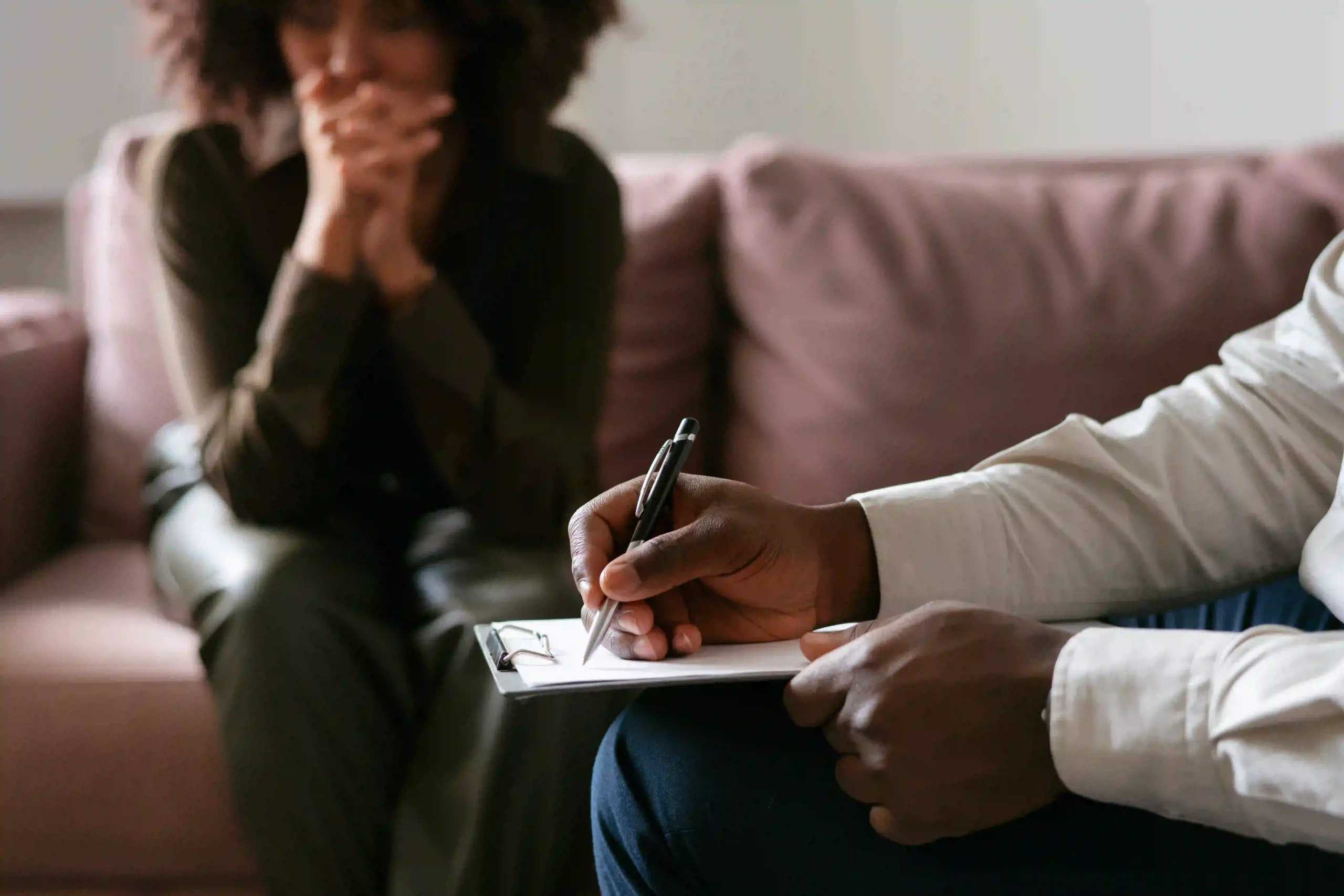 Close-up of a therapy session where a counselor writes notes on a clipboard while a person sits in the background with hands clasped, expressing emotional distress.