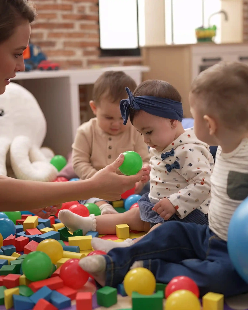 Preschool teacher playing with toddlers on the floor surrounded by colorful toys.