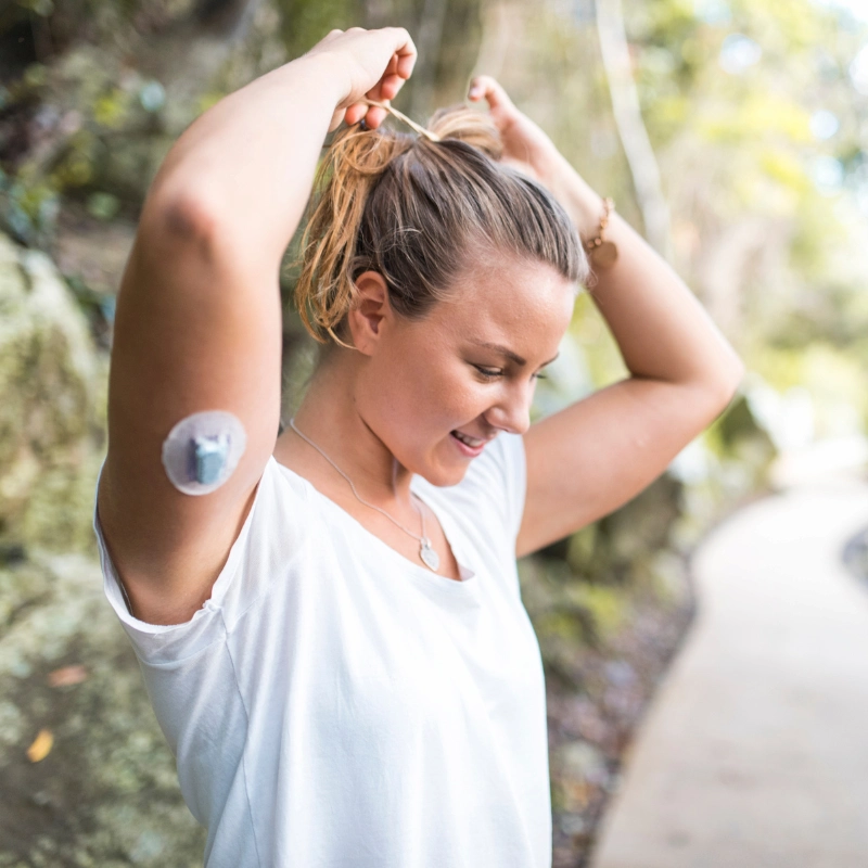Woman with a continuous glucose monitor sensor on her arm tying her hair outdoors.