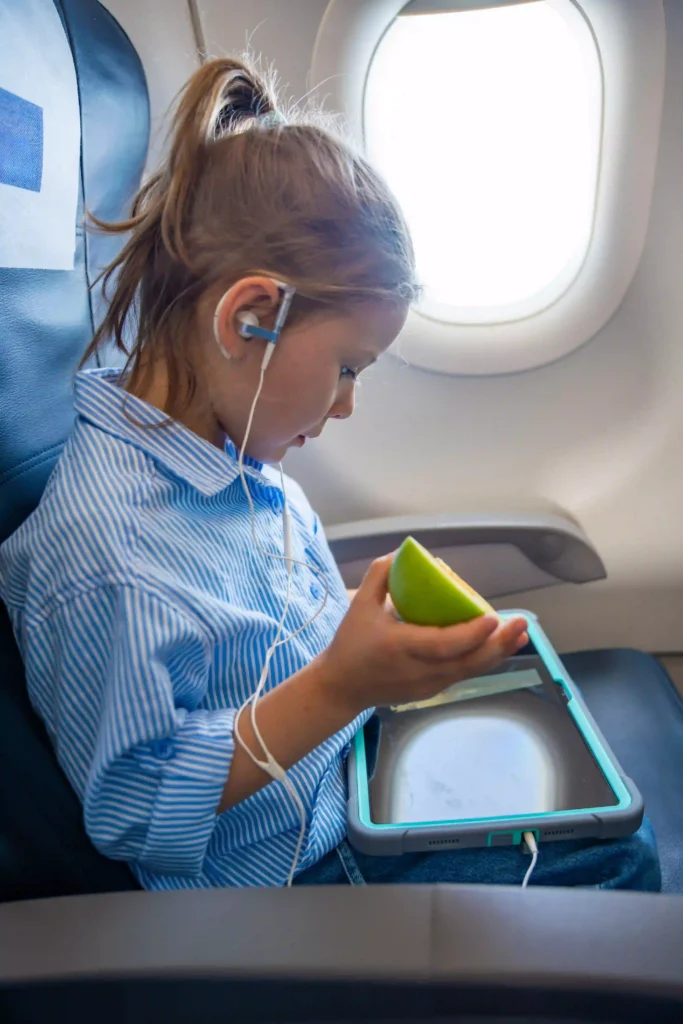 Child sitting on an airplane watching a tablet and eating a green apple during flight.