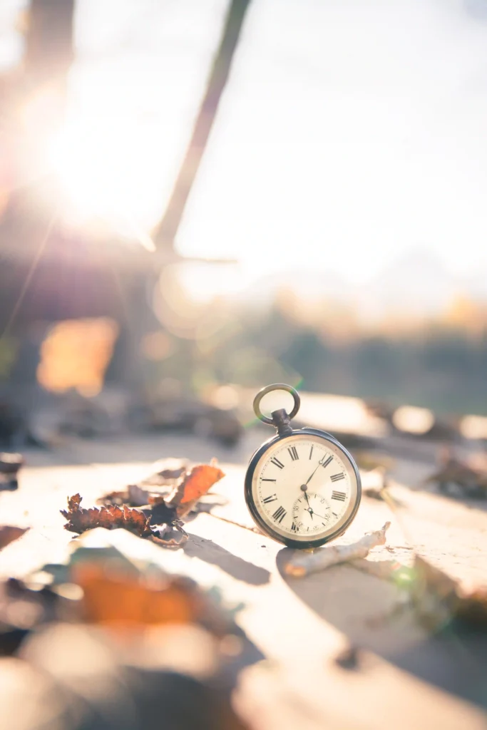 Vintage pocket watch on a wooden surface surrounded by autumn leaves in sunlight